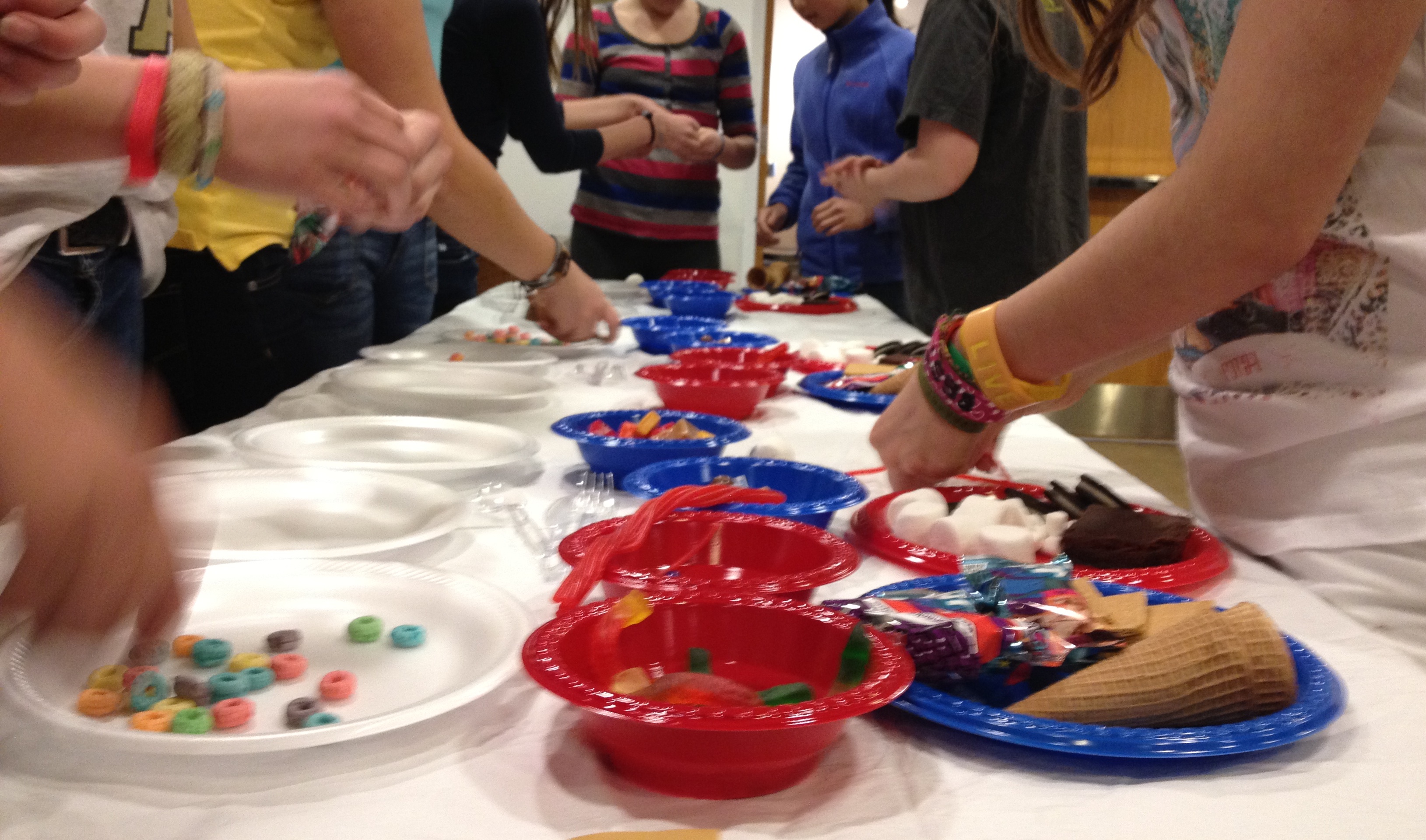 A photograph of teens from the shoulders down, gathered around a table covered in plates and bowls of candy and junk food, as they try to win a Teen Iron Chef competition